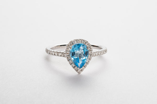 This white golden ring is set with diamonds on the side and a stunning Swiss blue Topaz. The Topaz is surrounded by a diamond halo for extra brilliance.   The Swiss Topaz is a vivid blue and gives your look a nice touch of color.  The pear cut is the mosted wanted fancy shape.   18kt white gold Total diamond weight for the ring approx. 0.23ct round brilliant diamonds Swiss blue Topaz dimensions 7.0 x 5.0mm